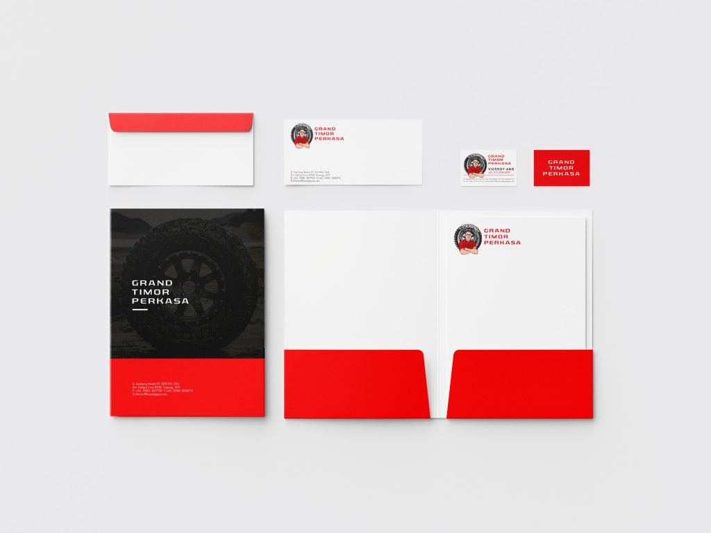Aplikasi desain logo Grand Timor Perkasa pada alat tulis kantor dan foto ban mobil. Terlihat desain untuk map folder (bagian dalam dan luar), kop surat, amplop, dan kartu nama. Logo Grand Timor Perkasa menampilkan siluet wajah pria berkacamata dalam lingkaran, dengan teks "GRAND TIMOR PERKASA" di samping kanan. Warna merah dan putih mendominasi desain. Di bagian kiri bawah, terdapat foto close-up ban mobil dengan background gurun pasir, memberikan kesan tangguh dan petualangan.